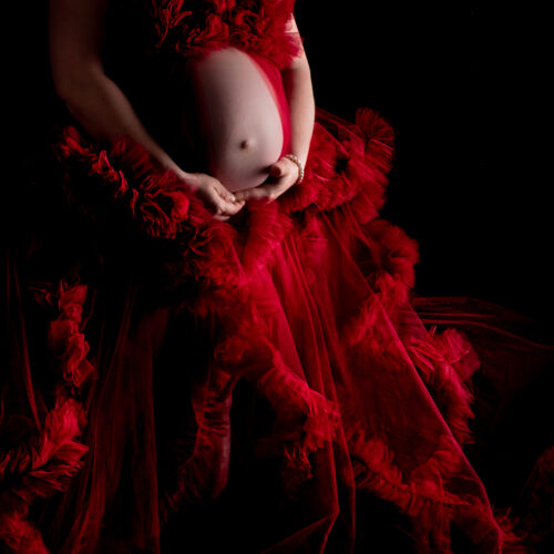 Ein rotes Tüllkleid umringt den Babybauch, welcher festgehalten wird von der Mutter beim Babybauch Foto Shooting.