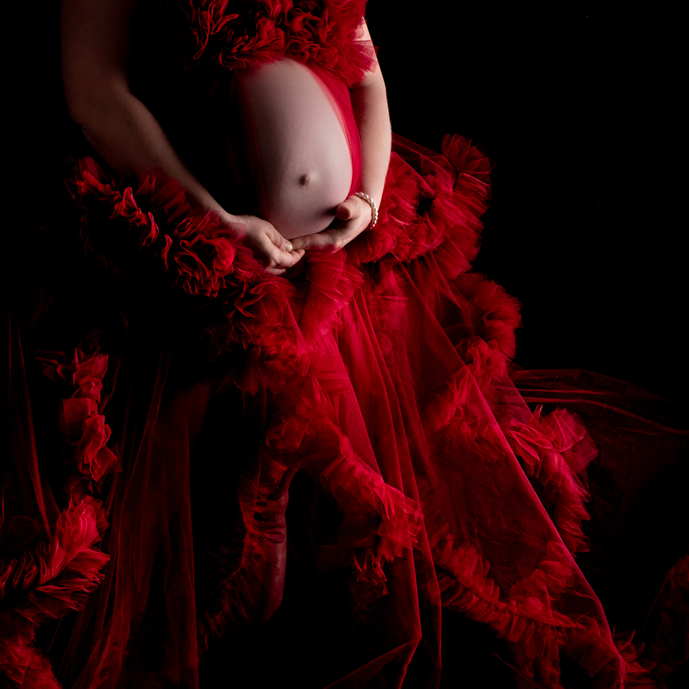 Ein rotes Tüllkleid umringt den Babybauch, welcher festgehalten wird von der Mutter beim Babybauch Foto Shooting.