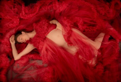 Schlafende Schönheit beim Babybauch Foto Shooting mit rotem Tüll umringt.