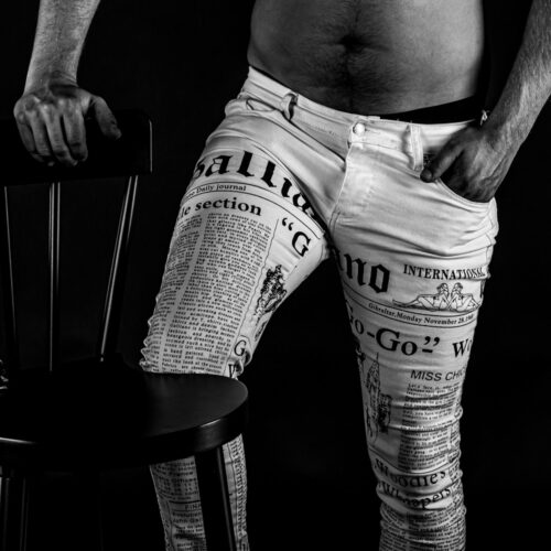 Eine schwarz weiss Aktfotografie eines Erotikshootings, wo der Herr mit weissen Textbedruckten Hosen bis zum Bauchnabel ersichtlich ist. er stütz sich mit der rechten Hand auf einen Sessel und hebt sein rechtes Bein.