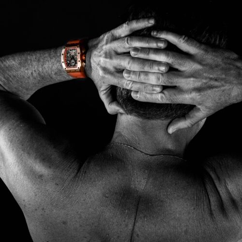 Zum Thema erlebe Erotikshootings mit Leidenschaft sehen wir hier ein männliches Model in schwarz-weiss mit dem Rücken zum Aktfotograf gedreht und seine Hände auf dem Hinterkopf verschränkt. Nur die Uhr auf seinem linken Arm ist in Farbe.