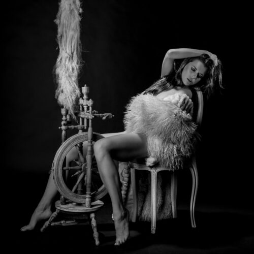 Beim Erotikfotoshooting sitzt die langhaarige Schönheit nach hinten gebeugt vor einem alten Spinnrad. Das erotische Foto ist in schwarz weiss gehalten.