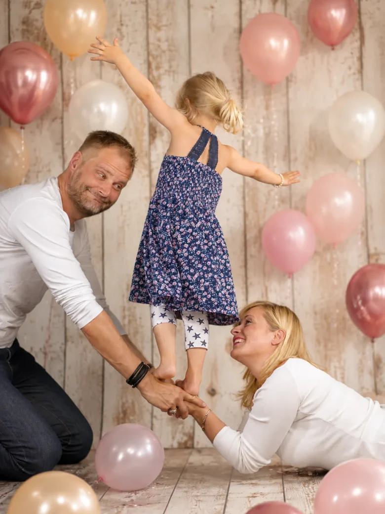 Kleines Mädchen im blauen Blumenkleid tanzt zwischen Vater und Mutter, ringsum Ballons in Rosé und Creme – Familienshooting Studio Hitzkirch Luzern