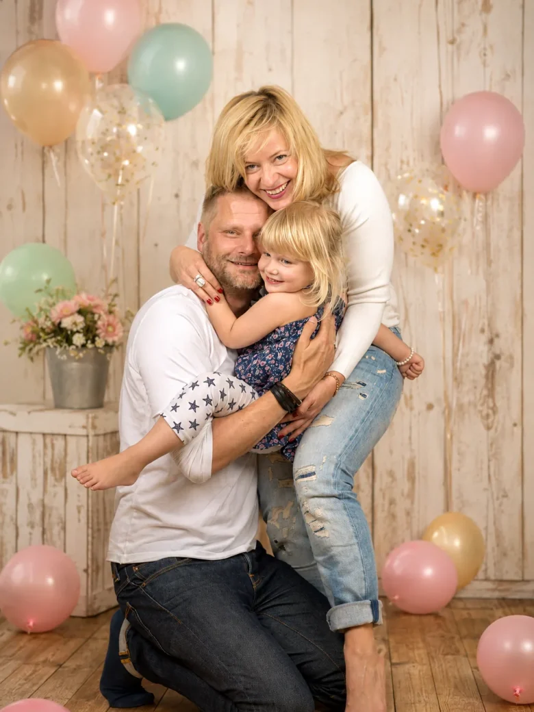 Vater kniet mit Mutter und Kleinkind auf dem Arm vor heller Holzwand mit Ballons und Blumen – Familienshooting mit Kind, Fotostudio Hitzkirch