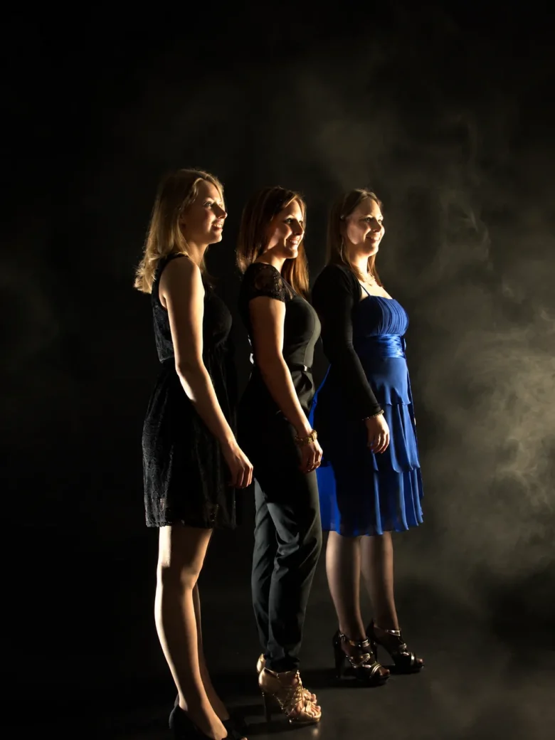 Drei junge Frauen in eleganten Cocktailkleidern stehen im Profil hintereinander im Nebellicht – Geschwistershooting im Fotostudio Hitzkirch