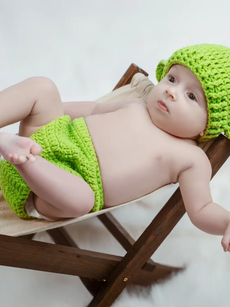 Fine-Art Newborn Fotografie – laechelndes Baby mit gruener Strickmuetze auf einem kleinen Holzstuhl vor weissem Hintergrund, Studio Hitzkirch Luzern