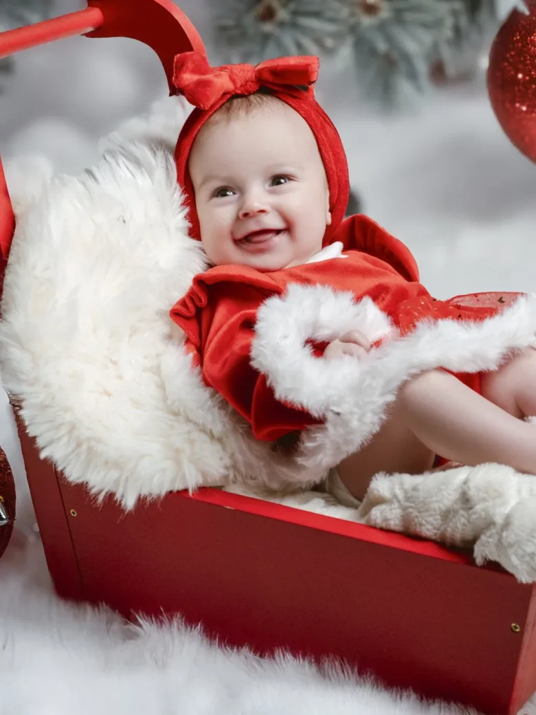 Fine-Art Newborn Fotografie – lachendes Baby im roten Weihnachtskleid in einem roten Holzschlitten mit Christbaumkugeln, Studio Hitzkirch Luzern